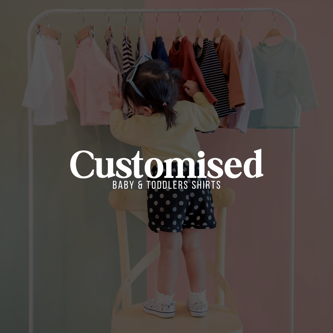Young child standing on a chair reaching for colourful baby and toddler shirts on a clothing rack, with bold text overlay reading "Customised Baby & Toddlers Shirts"