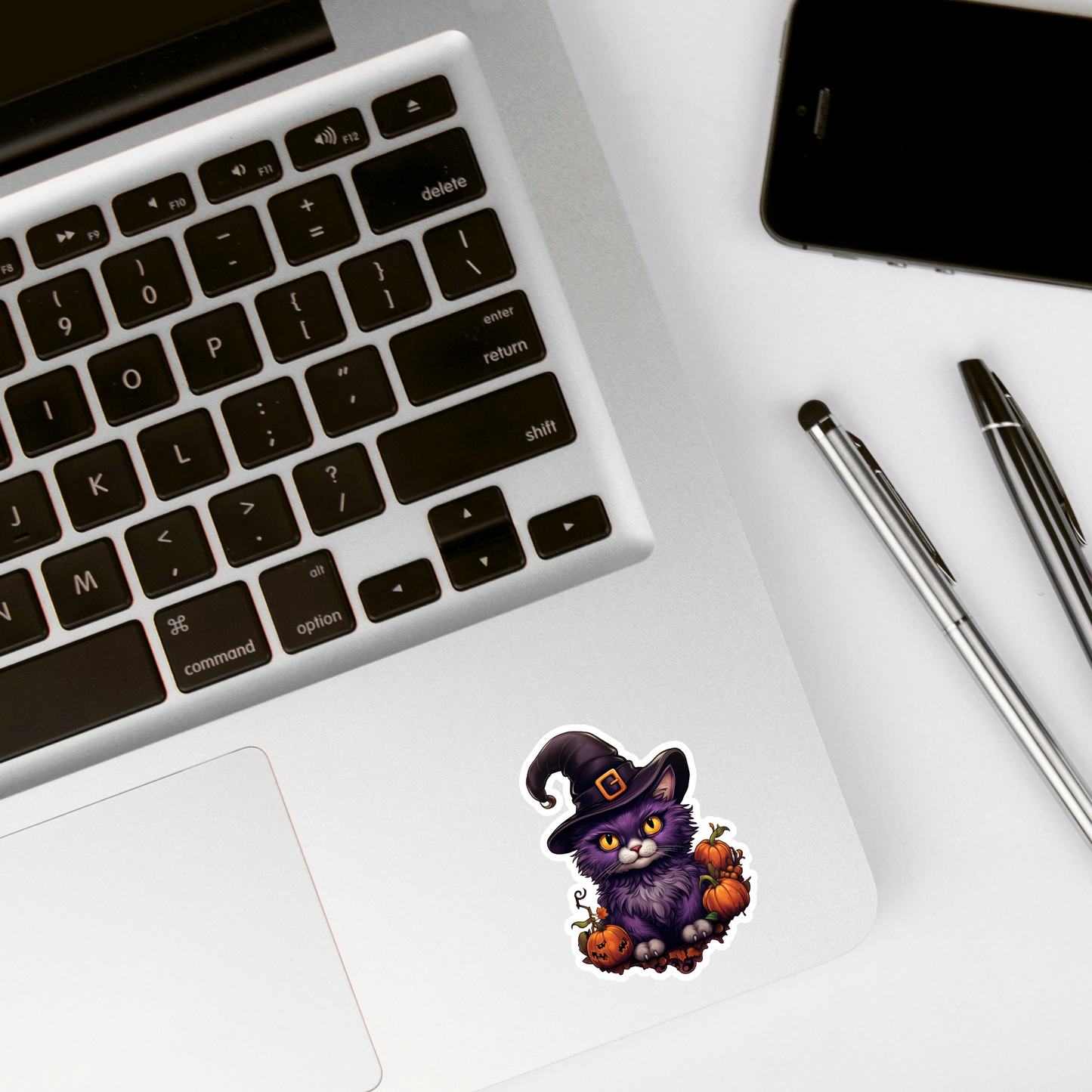 Halloween-themed sticker of a cat in a witch hat on a laptop with pens and a phone.