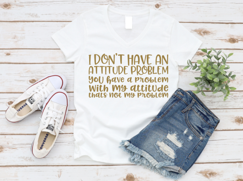 White V-neck t-shirt with gold text that reads “I don’t have an attitude problem. You have a problem with my attitude—that’s not my problem,” styled with ripped denim shorts, white Converse sneakers, and a small plant on a wooden background.
