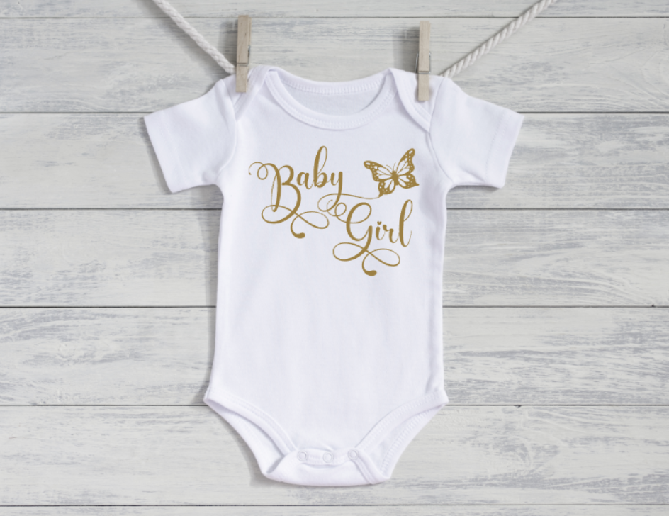 White baby onesie with 'Baby Girl' and butterfly design in gold on a wooden background