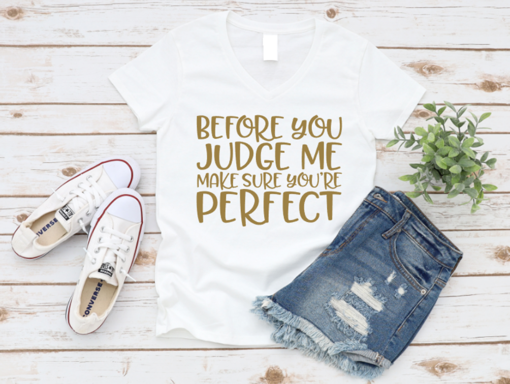 White t-shirt with gold text reading “Before you judge me make sure you’re perfect,” paired with denim shorts and white sneakers on a light wooden background.