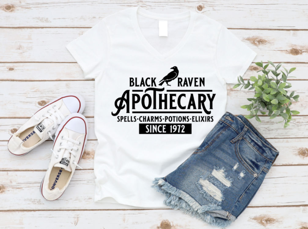 White t-shirt with “Black Raven Apothecary – Spells, Charms, Potions, Elixirs – Since 1972” printed in white. Styled with ripped denim shorts, white sneakers, and greenery on a white wooden backdrop.