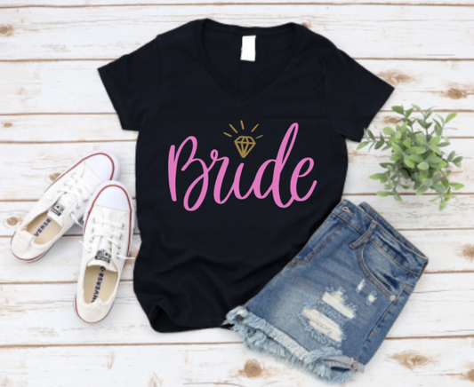 Black t-shirt with “Bride” in pink script and gold diamond icon, styled with distressed denim shorts and white Converse sneakers on a white wood background.