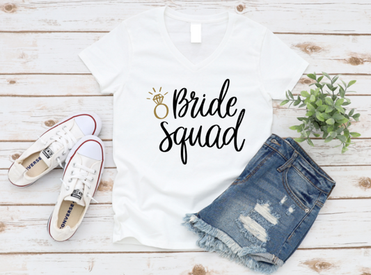 White t-shirt with “Bride Squad” text and gold ring graphic, styled with white sneakers and ripped denim shorts on a white wooden background.