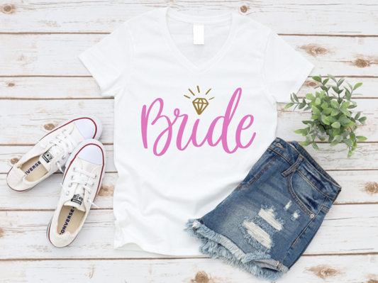 White t-shirt with “Bride” in pink script and gold diamond icon, styled with distressed denim shorts and white Converse sneakers on a white wood background.