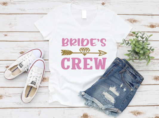 White t-shirt with “Bride’s Crew” pink and gold text, styled with ripped denim shorts and white sneakers on a wooden background.