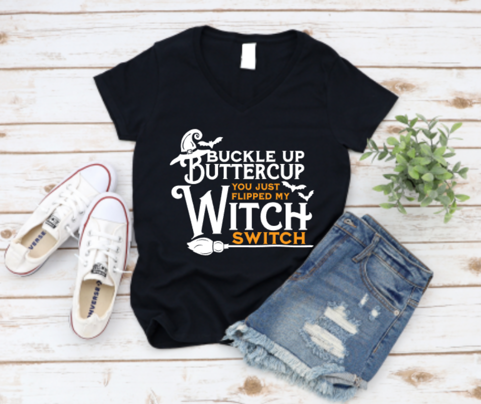Black t-shirt with "Buckle Up Buttercup – You Just Flipped My Witch Switch" text in black and orange, styled with ripped denim shorts and white sneakers on a white wood background.