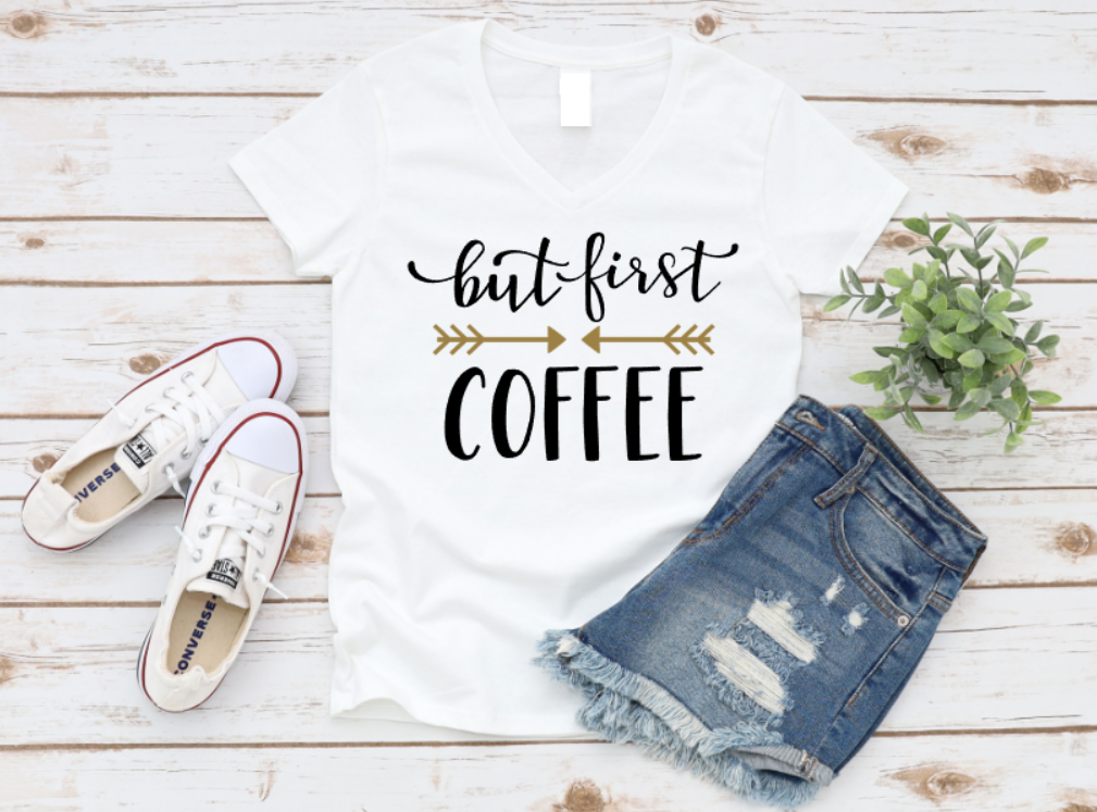 White t-shirt with "But First, Coffee" text in white and gold, styled with ripped denim shorts, white sneakers, and a plant on a wooden background.