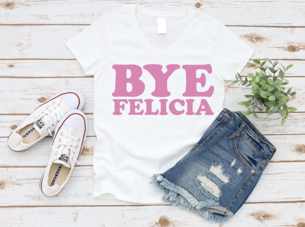 White t-shirt with bold pink “Bye Felicia” text, styled with denim shorts, sneakers, and a green plant on a wooden background.