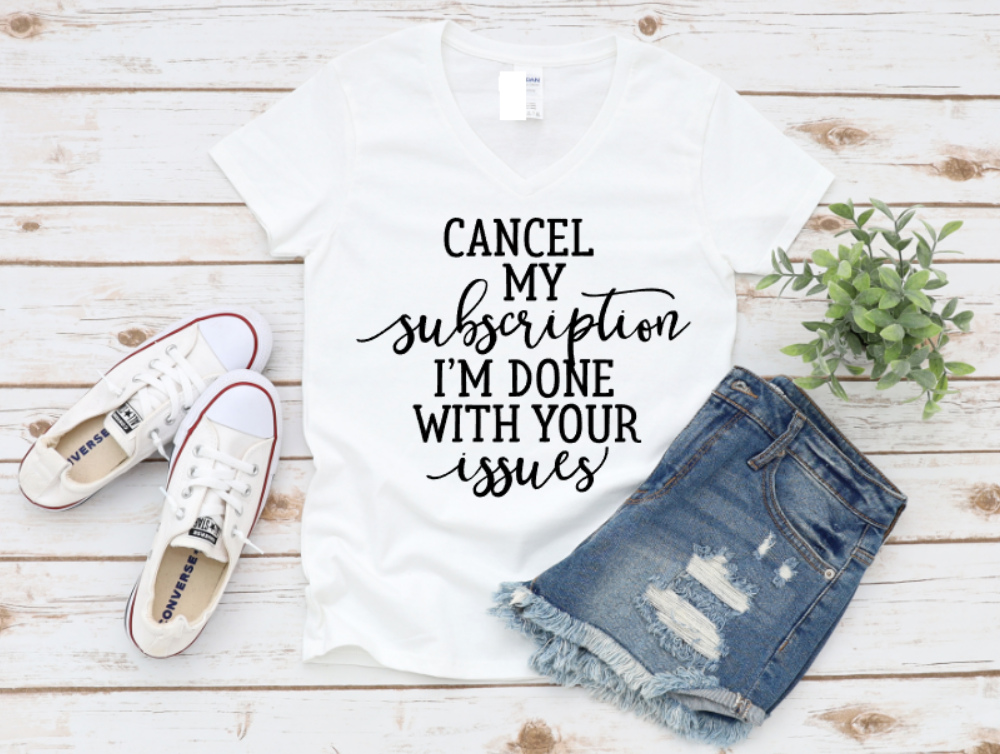 White t-shirt with white “Cancel my subscription I’m done with your issues” text, styled with distressed denim shorts, white sneakers, and greenery on a wooden background.