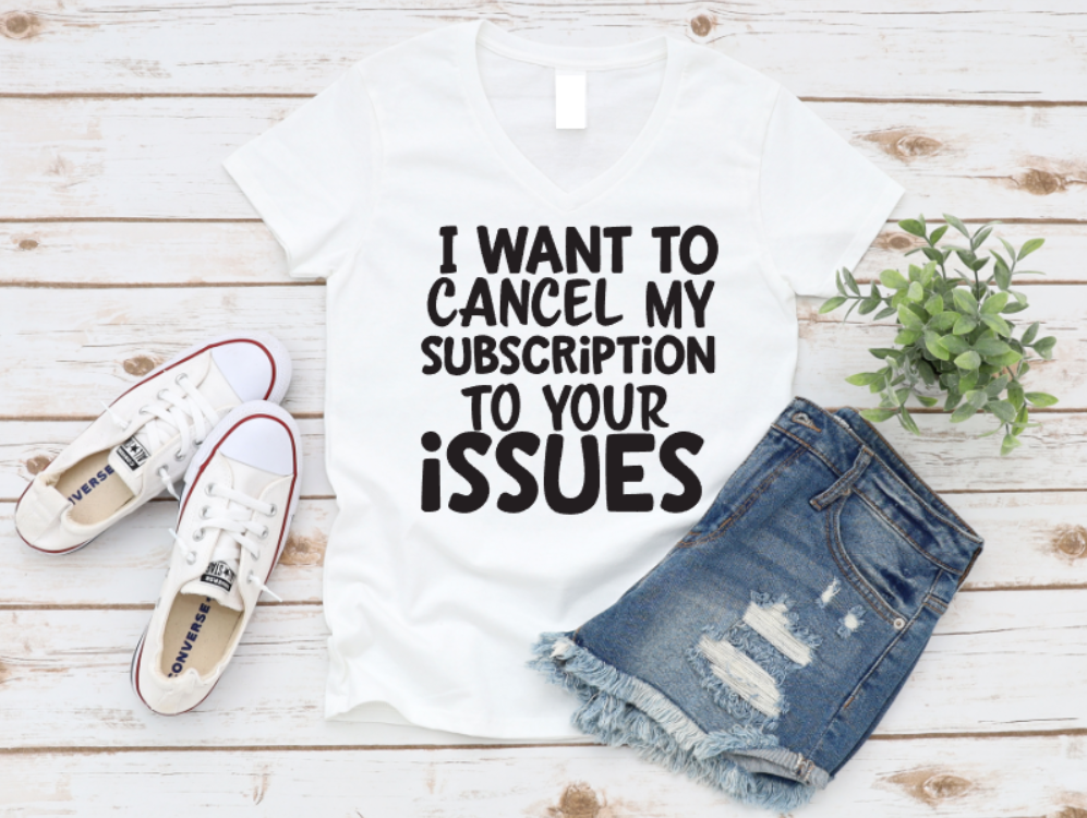 White t-shirt with the text “I want to cancel my subscription to your issues,” styled with ripped denim shorts, white sneakers, and a green plant on a wooden background.