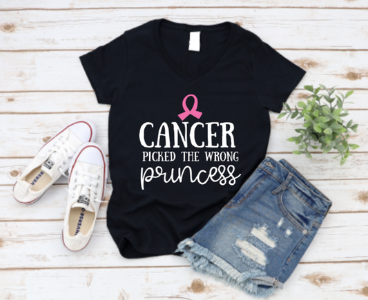 Black t-shirt with “Cancer Picked the Wrong Princess” text and pink ribbon, styled with denim shorts and white sneakers on a wooden surface.