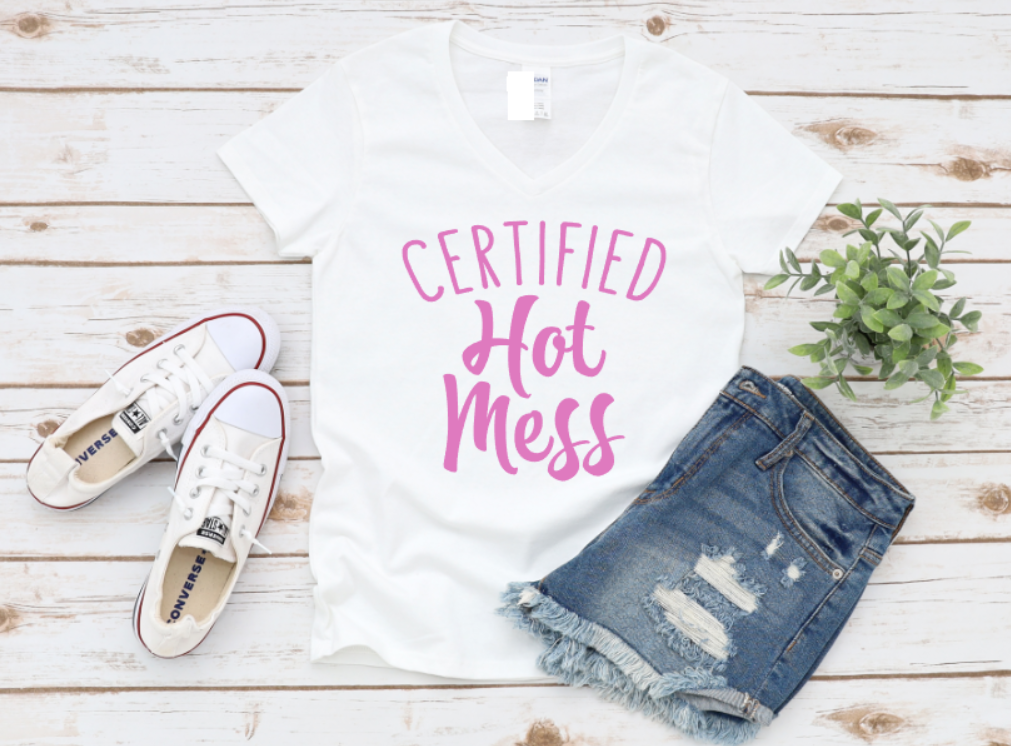 White t-shirt with "Certified Hot Mess" text in pink, styled with white sneakers and distressed denim shorts on a wooden background.