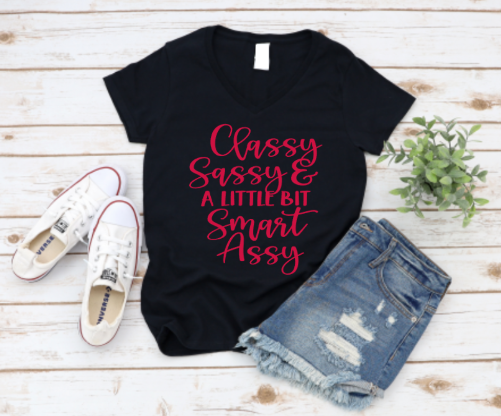 Black t-shirt with “Classy Sassy & a Little Bit Smart Assy” red text, styled with denim shorts and white sneakers on a light wooden surface.