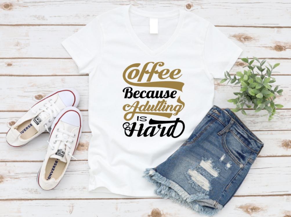 White t-shirt with "Coffee Because Adulting Is Hard" design in brown, gold, and black lettering, styled with ripped denim shorts and white Converse sneakers on a wooden background.