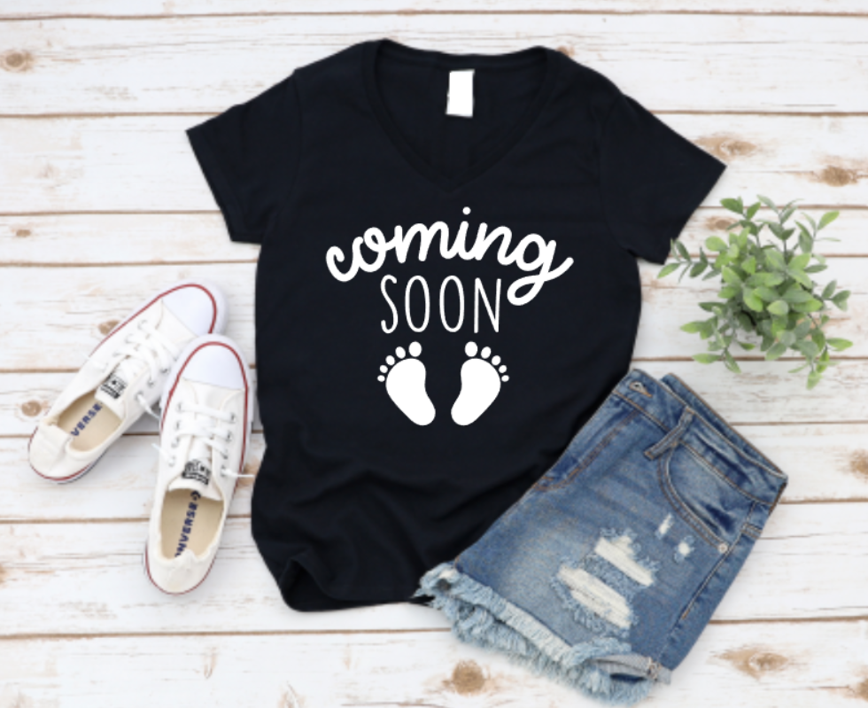 Black maternity t-shirt with the phrase “Coming Soon” and black baby footprints, styled flat lay with white sneakers and ripped denim shorts on a wooden background.