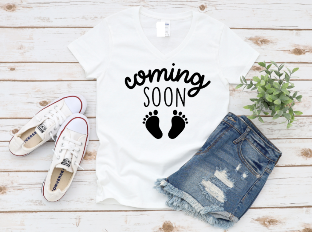 White maternity t-shirt with the phrase “Coming Soon” and black baby footprints, styled flat lay with white sneakers and ripped denim shorts on a wooden background.