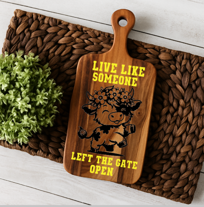 Wooden paddle-style charcuterie board featuring a black floral cow graphic and the phrase “Live Like Someone Left the Gate Open” in bold yellow vinyl. The board rests on a woven placemat with a green faux plant nearby, styled in a rustic farmhouse setting.