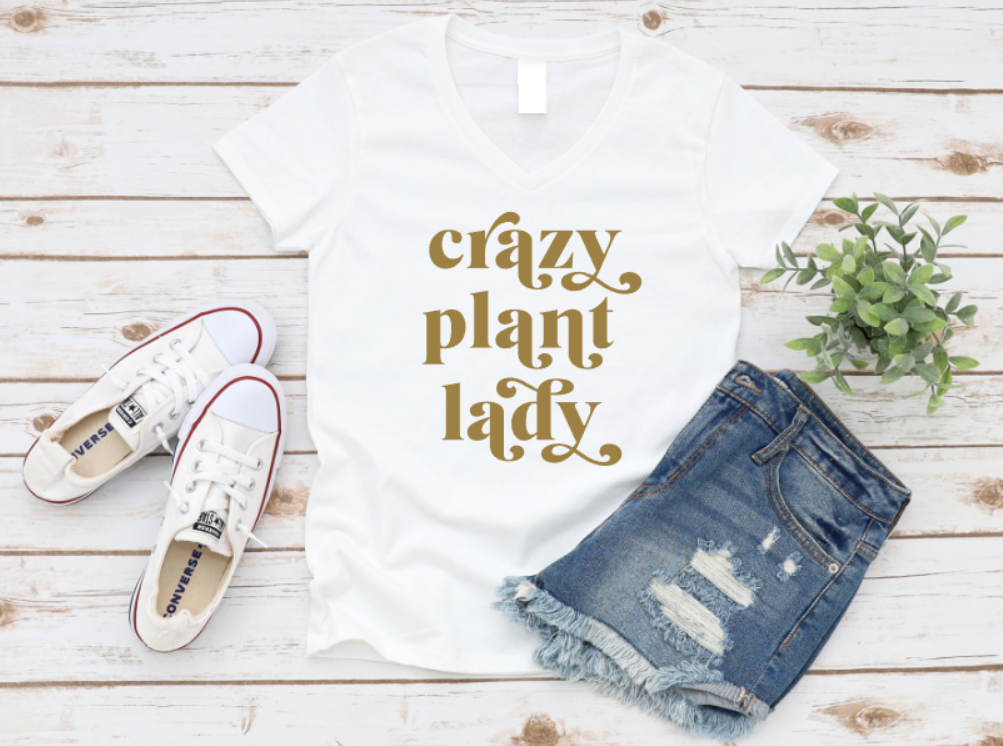 White t-shirt with "Crazy Plant Lady" text in gold script, styled with distressed denim shorts and white Converse sneakers on a wooden background.