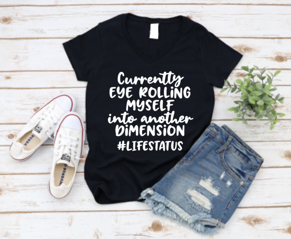 Black t-shirt laid flat with white bold text reading “Currently Eye Rolling Myself Into Another Dimension #LIFESTATUS”, styled with ripped denim shorts and white Converse shoes on a wooden background