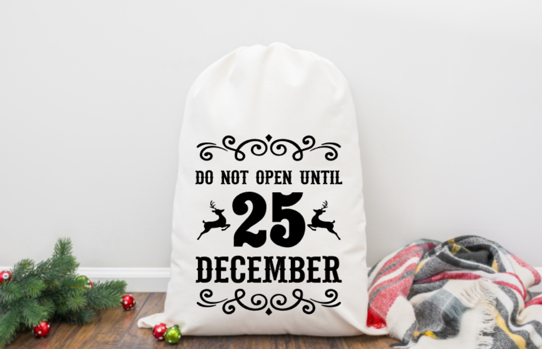 White sack with 'Do not open until 25 December' text on a wooden surface with Christmas decorations.
