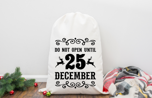 White sack with 'Do not open until 25 December' text on a wooden surface with Christmas decorations.