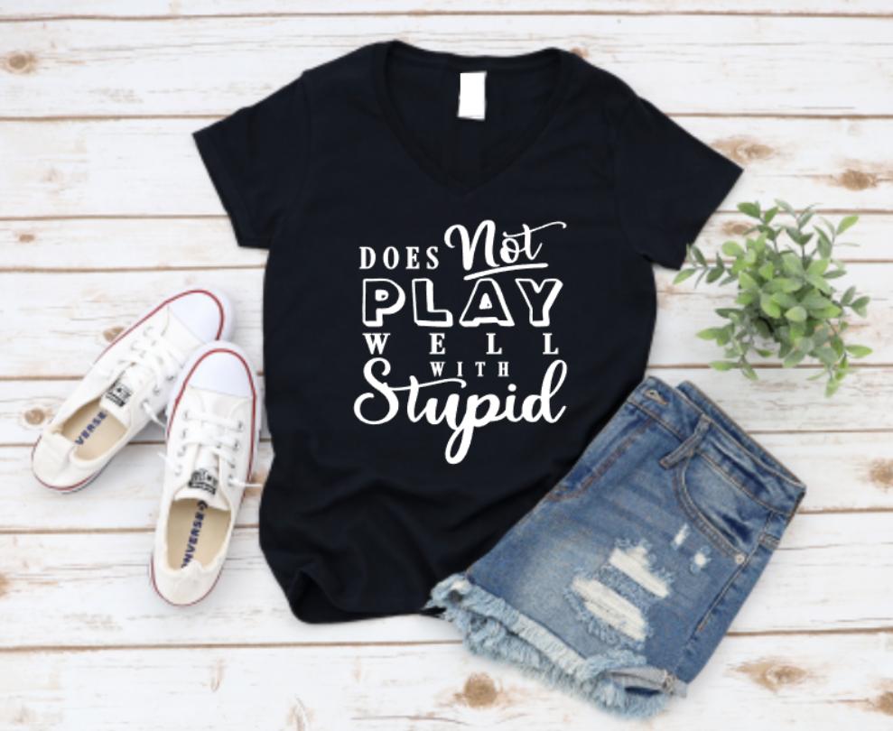 Flat lay of a black v-neck t-shirt with the phrase “Does Not Play Well With Stupid” in bold white lettering, styled with denim shorts, Converse sneakers, and a plant on a wooden background.