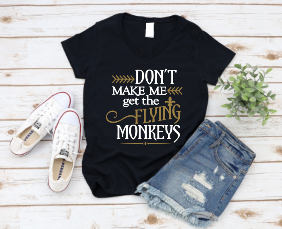 Flat lay of a black t-shirt with the phrase “Don’t Make Me Get the Flying Monkeys” in bold white and gold lettering, styled with ripped denim shorts, Converse sneakers, and a green plant on a wooden background.