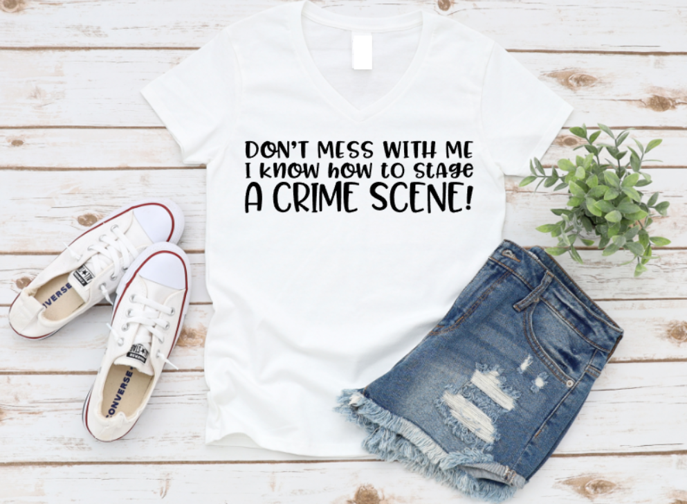 White v-neck t-shirt with the phrase “Don’t mess with me I know how to stage a crime scene” in bold white text, styled with denim shorts and white Converse sneakers on a rustic wood background.