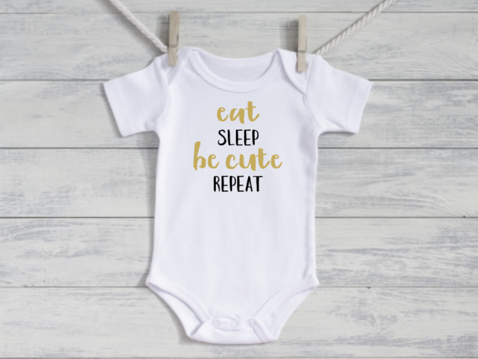 White baby onesie with gold text 'eat sleep be cute repeat' on a wooden background