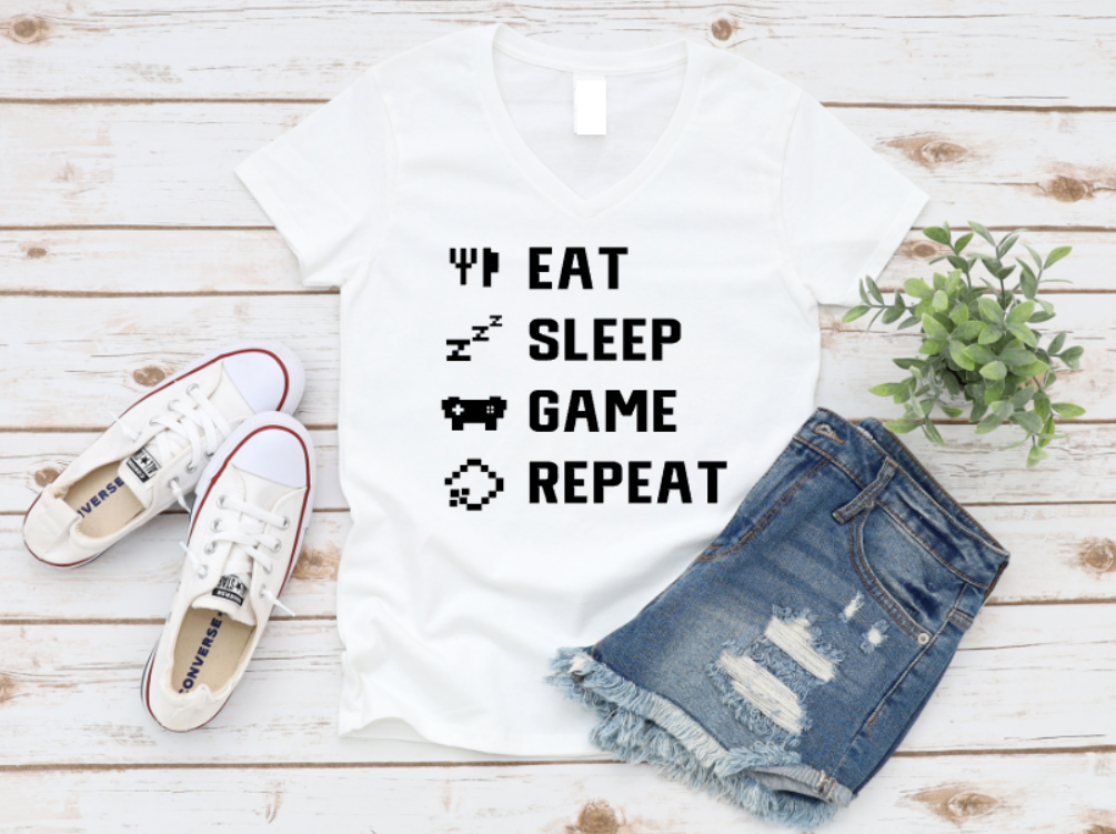 White t-shirt with white pixel-style text reading "Eat Sleep Game Repeat" alongside gaming and sleep icons, perfect for gamers.