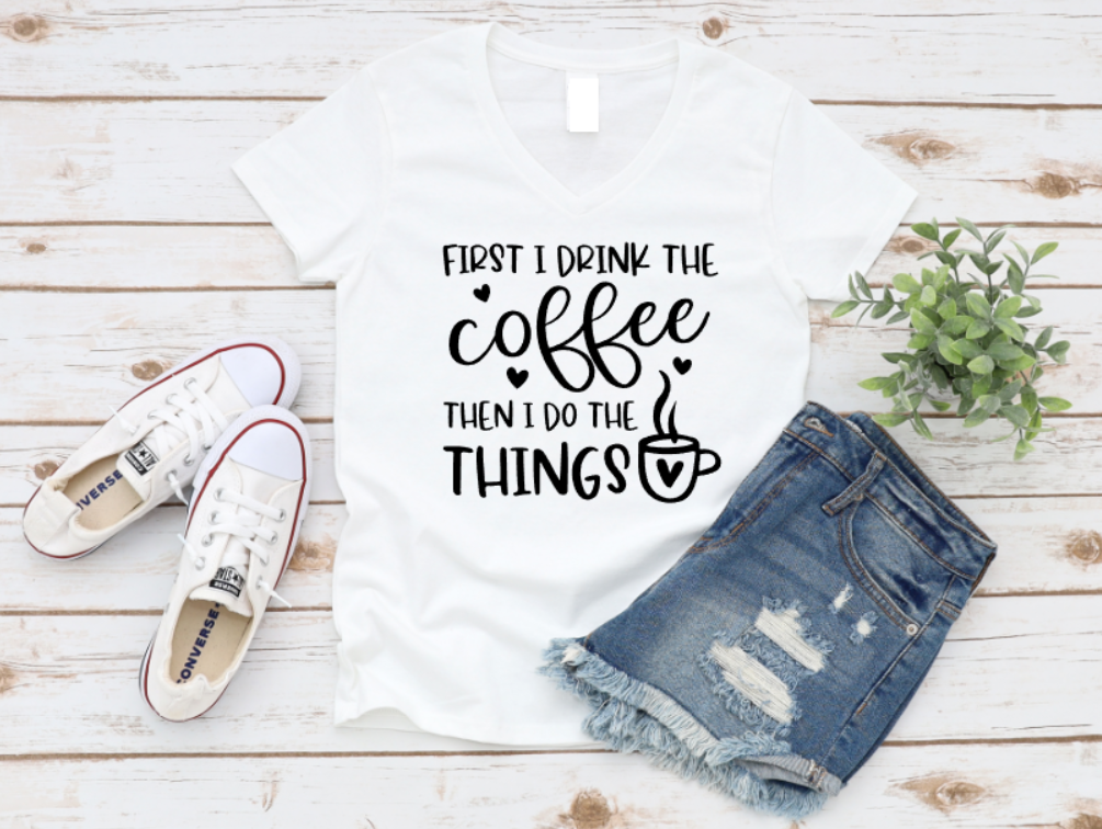 White t-shirt with the phrase "First I Drink the Coffee Then I Do the Things" in playful black font, styled with denim shorts and white Converse sneakers on a wooden background.