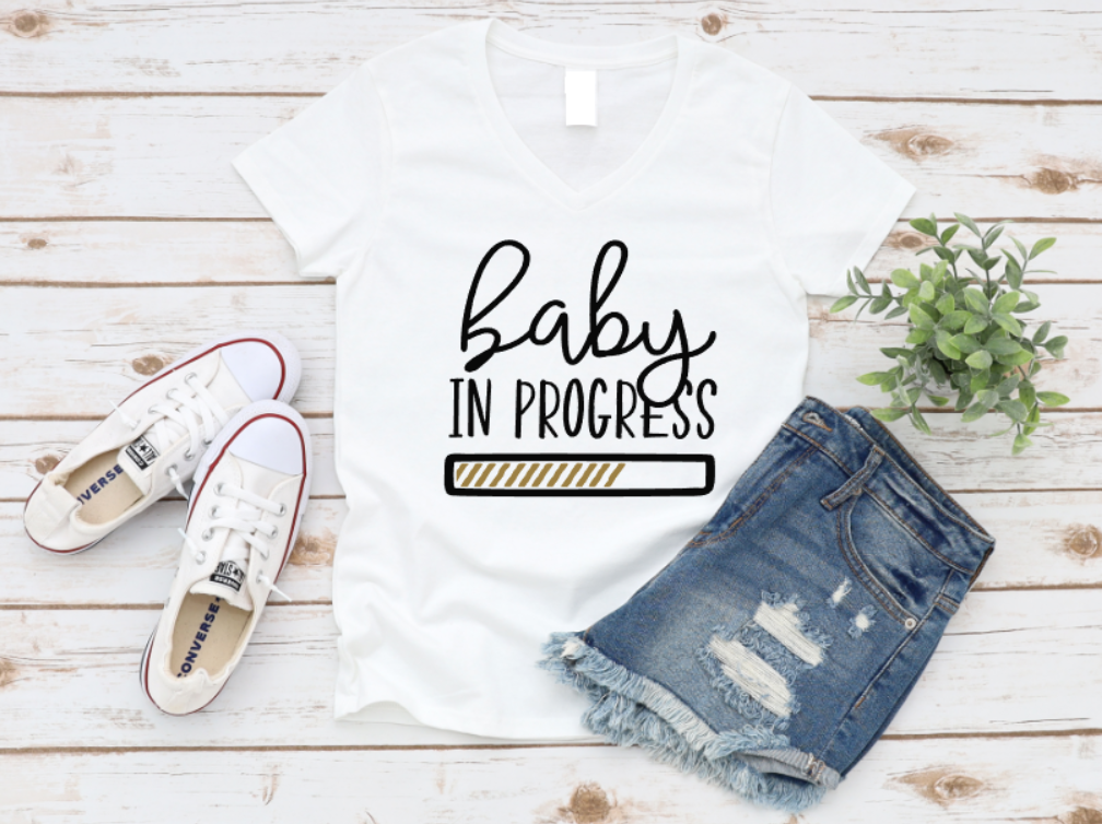 White t-shirt with "baby in progress" text and a yellow progress bar graphic, styled with white sneakers and ripped denim shorts on a wooden background.