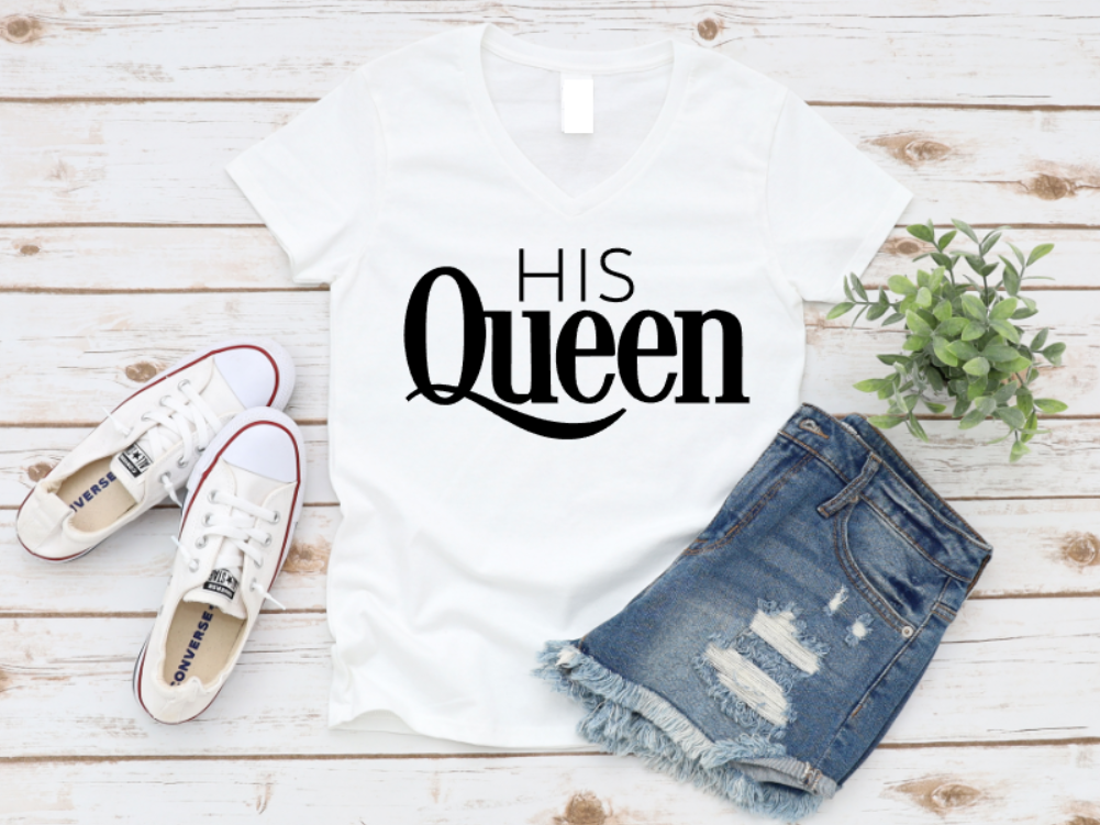 White top with 'His Queen' text, white sneakers, and denim shorts on a wooden surface.