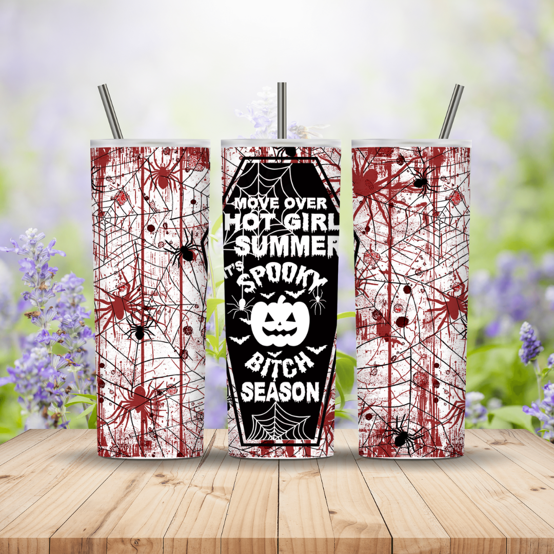 Three Halloween-themed tumblers with spider webs and text on a wooden surface with a blurred floral background.