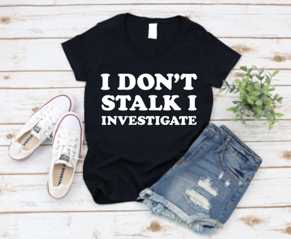 Black t-shirt with 'I Don't Stalk I Investigate' text, paired with denim shorts and white sneakers on a wooden surface.