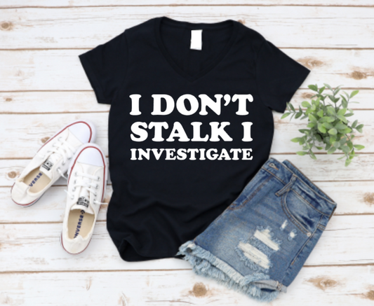 Black t-shirt with 'I Don't Stalk I Investigate' text, paired with denim shorts and white sneakers on a wooden surface.
