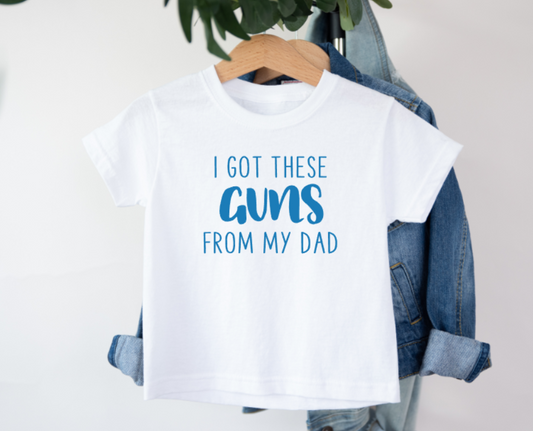 White t-shirt with 'I got these guns from my dad' text on a hanger with denim jacket.