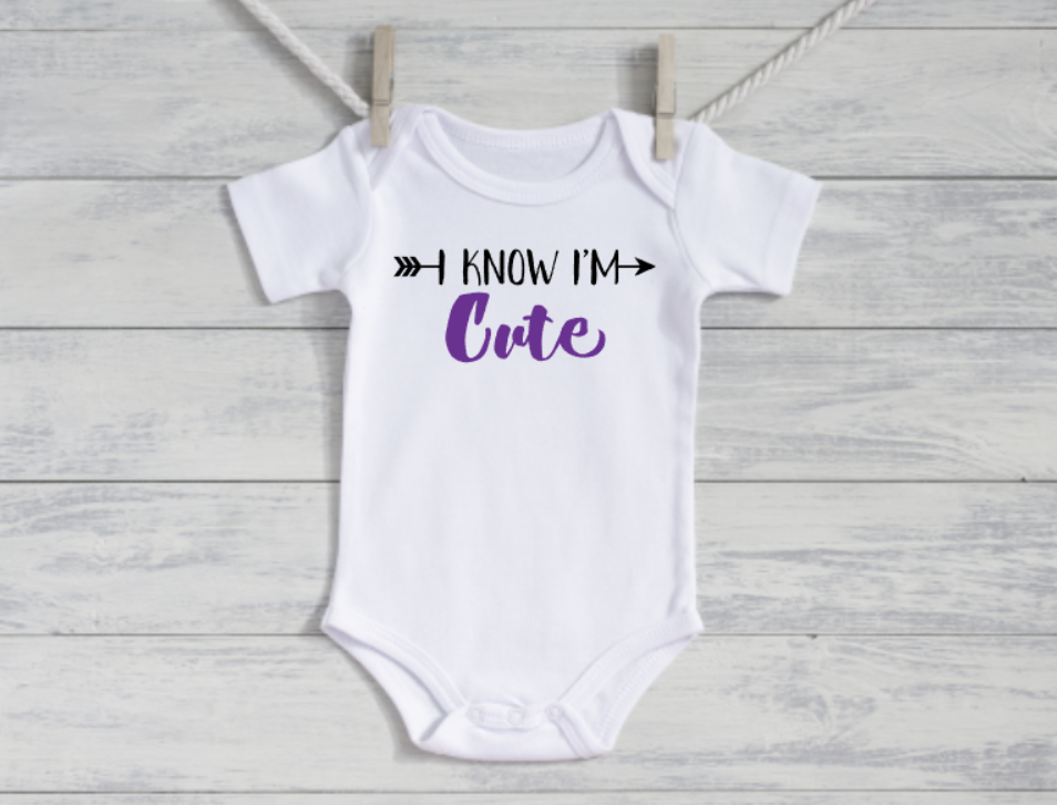 White baby onesie with 'I Know I'm Cute' text on a wooden surface