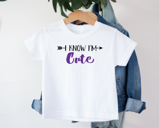 White t-shirt with 'I Know I'm Cute' text on a hanger with denim jacket.
