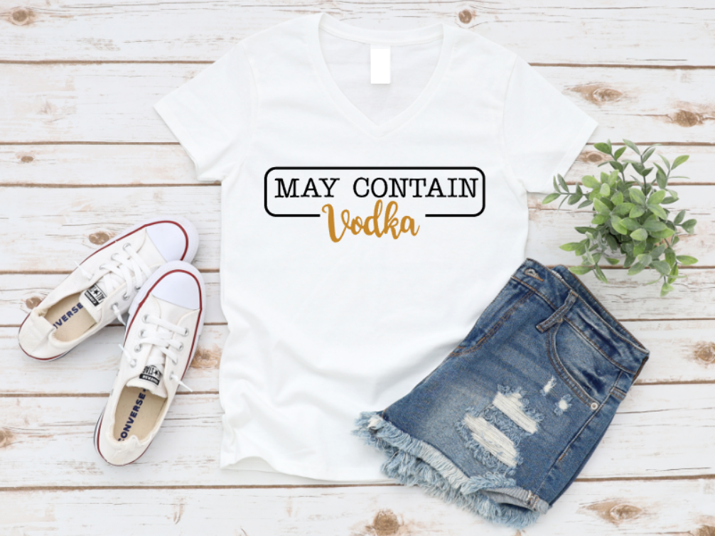 Flat lay of a white t-shirt with the phrase “May Contain Vodka” in black and gold text, styled with denim shorts and white Converse sneakers on a wooden background.