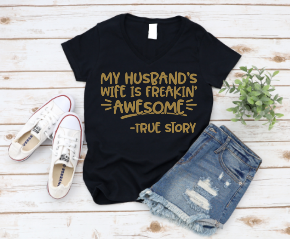 Black t-shirt with gold text 'My husband's wife is freakin' awesome - true story' on a wooden surface with shoes and shorts. www.missmafia.com