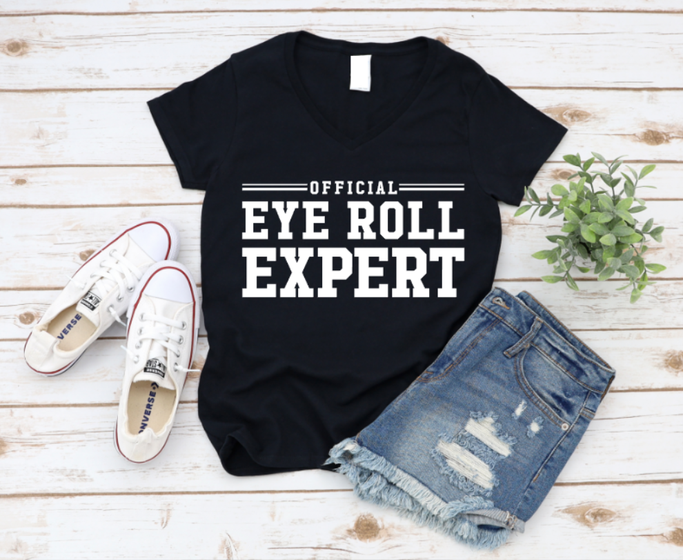 Flat lay of a Black t-shirt with bold black text reading “Official Eye Roll Expert,” styled with denim shorts, Converse sneakers, and a plant on a rustic wooden background.