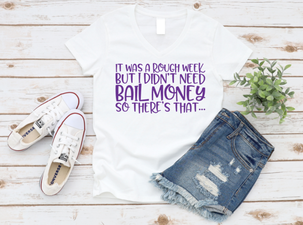 White t-shirt with purple "It was a rough week but I didn’t need bail money, so there’s that..." text, styled with denim shorts and white sneakers on a wooden background.