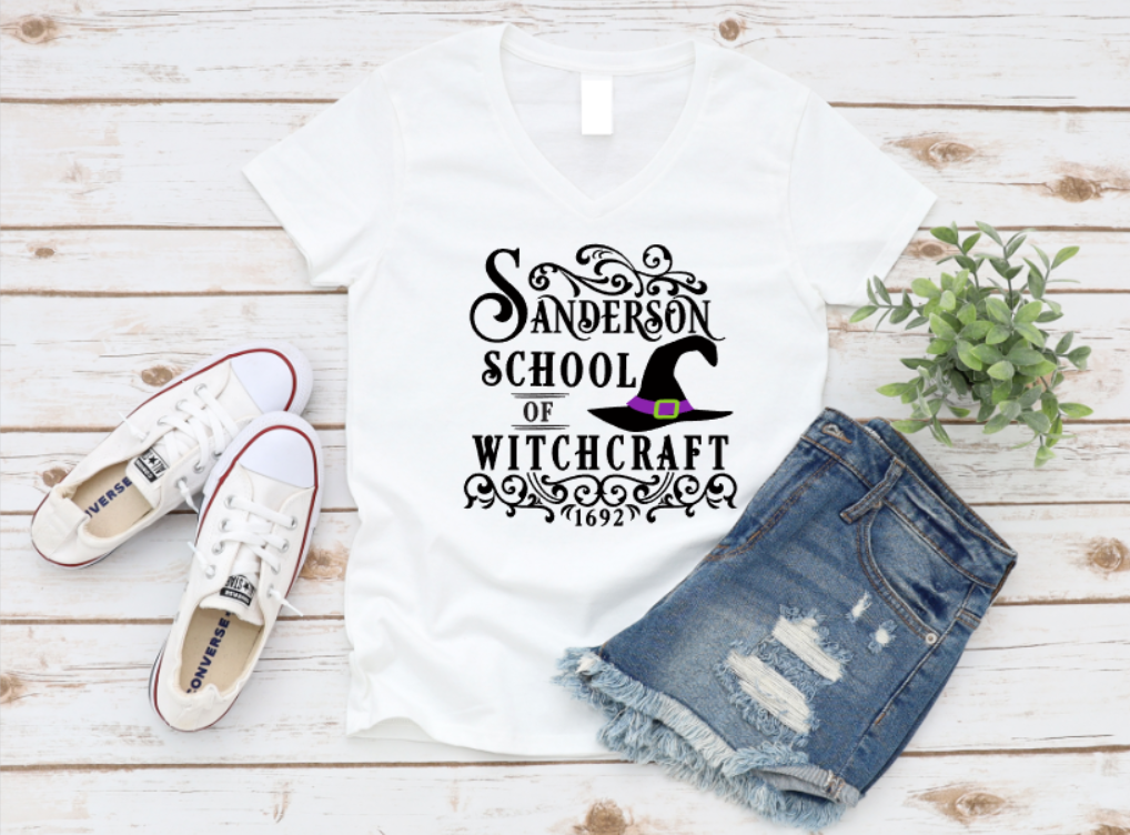 White t-shirt with white gothic-style text that reads "Sanderson School of Witchcraft – 1692" and features a purple and green witch hat illustration. Shirt is styled flat lay with denim shorts, white Converse sneakers, and a small green plant on a light wood background. www.missmafia.com