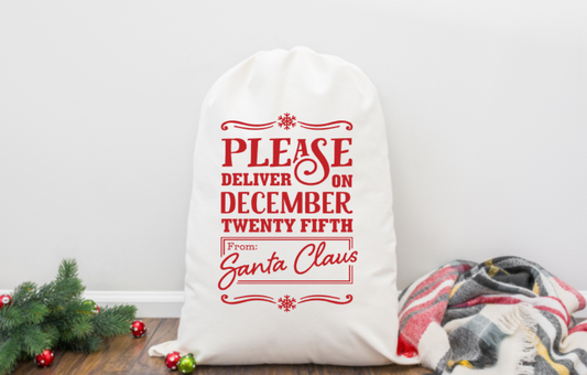 White sack with red text 'Please deliver on December twenty fifth from Santa Claus' on a wooden floor with Christmas decorations.