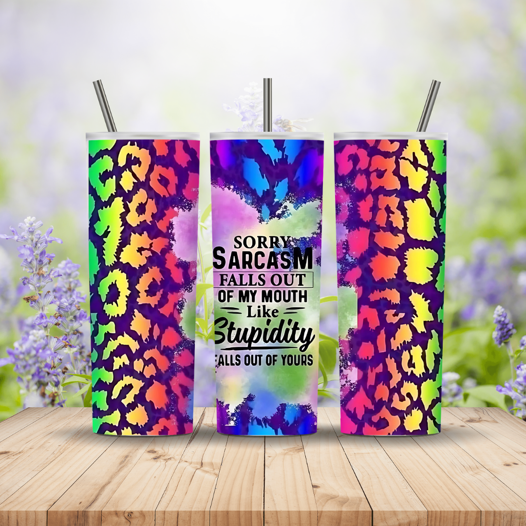 Three colorful tumblers with leopard print designs and a humorous quote on a wooden surface with a blurred floral background.