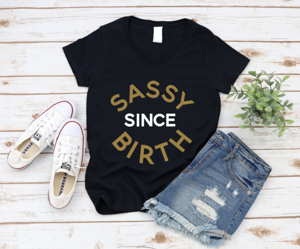 Flat lay of a black t-shirt with the phrase “Sassy Since Birth” printed in bold gold and white text, styled with distressed denim shorts, white Converse sneakers, and a small green plant on a white wooden background. | www.missmafia.com