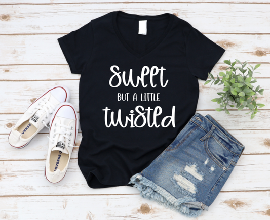 Flat lay of a black t-shirt with the phrase “Sweet But a Little Twisted” in white text, styled with ripped denim shorts, white Converse sneakers, and a small green plant on a white wooden background. www.missmafia.com