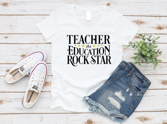 White t-shirt with 'Teacher aka Education Rock Star' text, white sneakers, and denim shorts on a wooden surface. www.missmafia.com